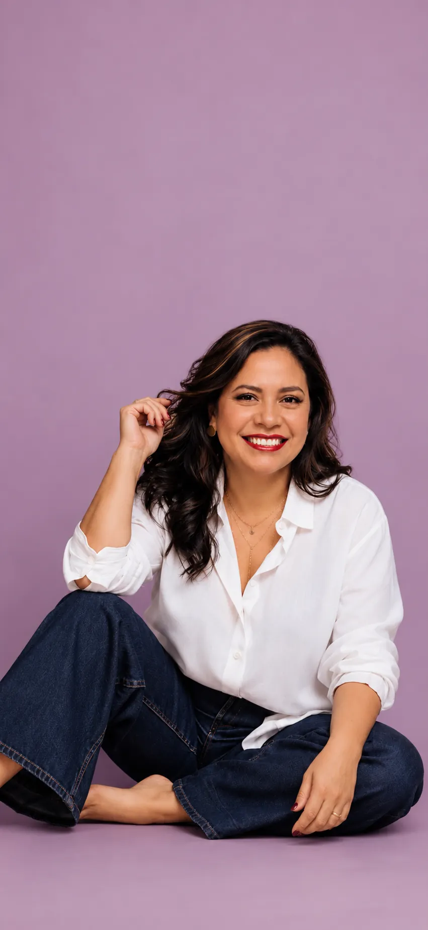 Ana Casalegno sonriendo, sentada en el suelo con camisa blanca y jeans sobre fondo lila